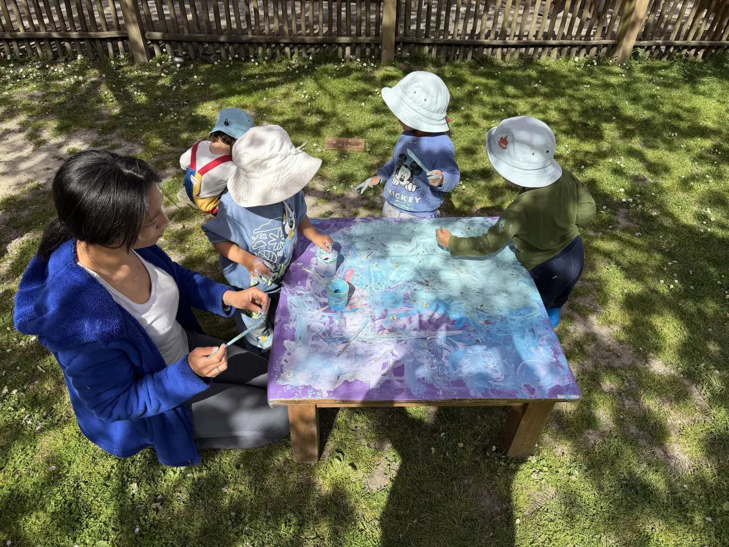 A teacher and children painting together at an outdoor table