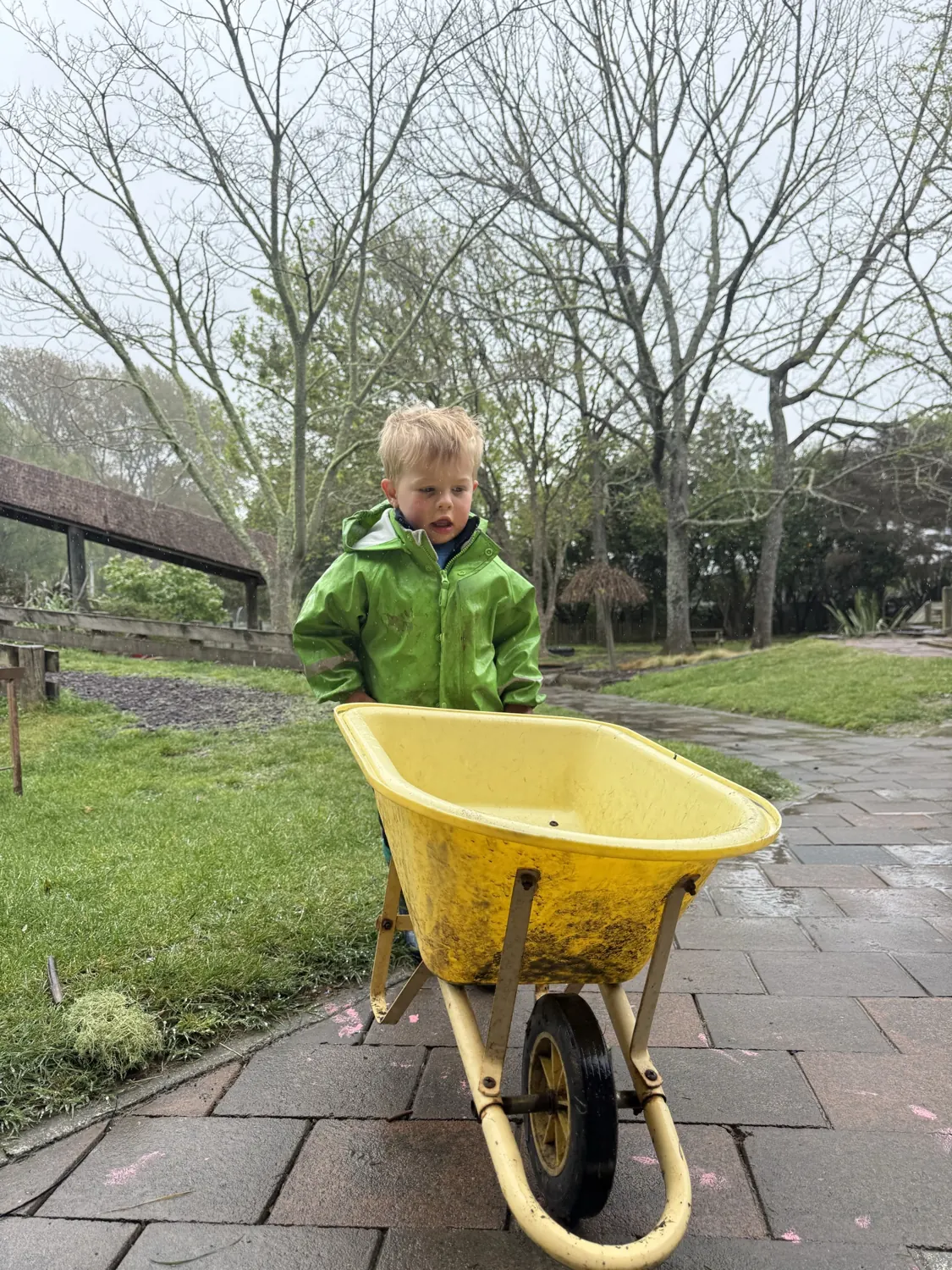 A child in a raincoat pushing a yellow wheelbarrow along the path