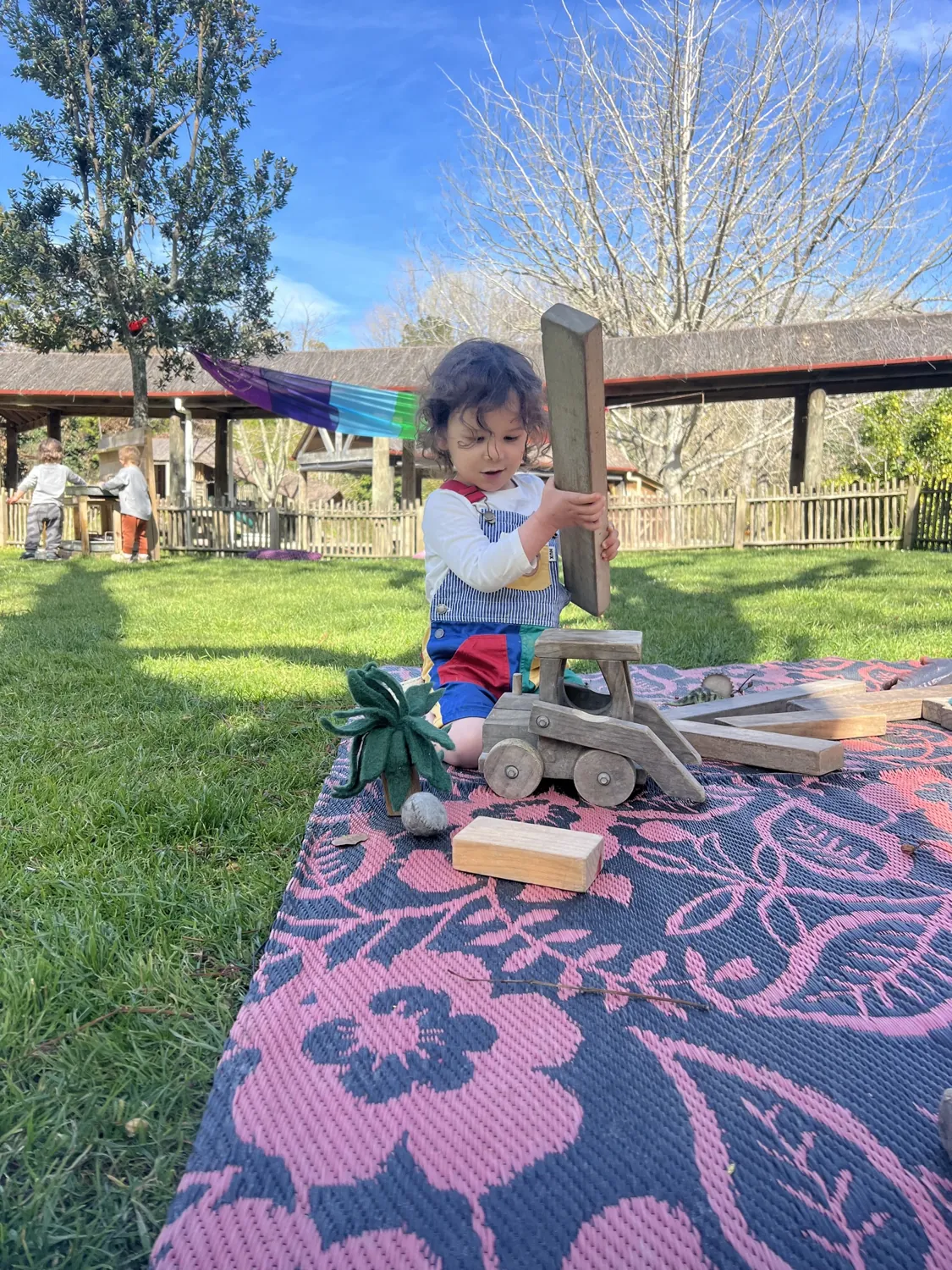 A toddler building with wooden blocks on a rug on the lawn, with the verandah and trees behind
