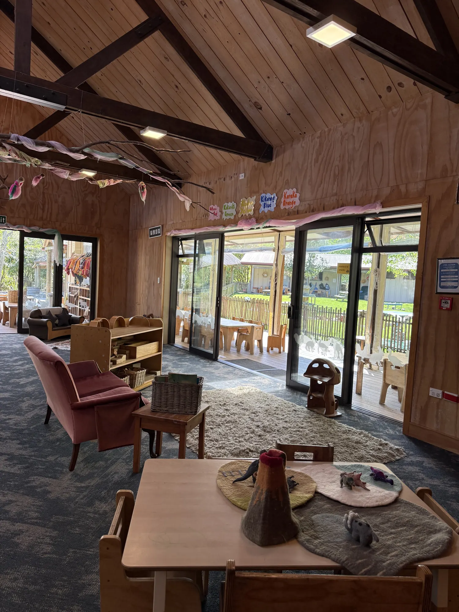 A timber-lined room with a high beamed ceiling, a rug, armchair and low tables, with glass doors opening to the deck