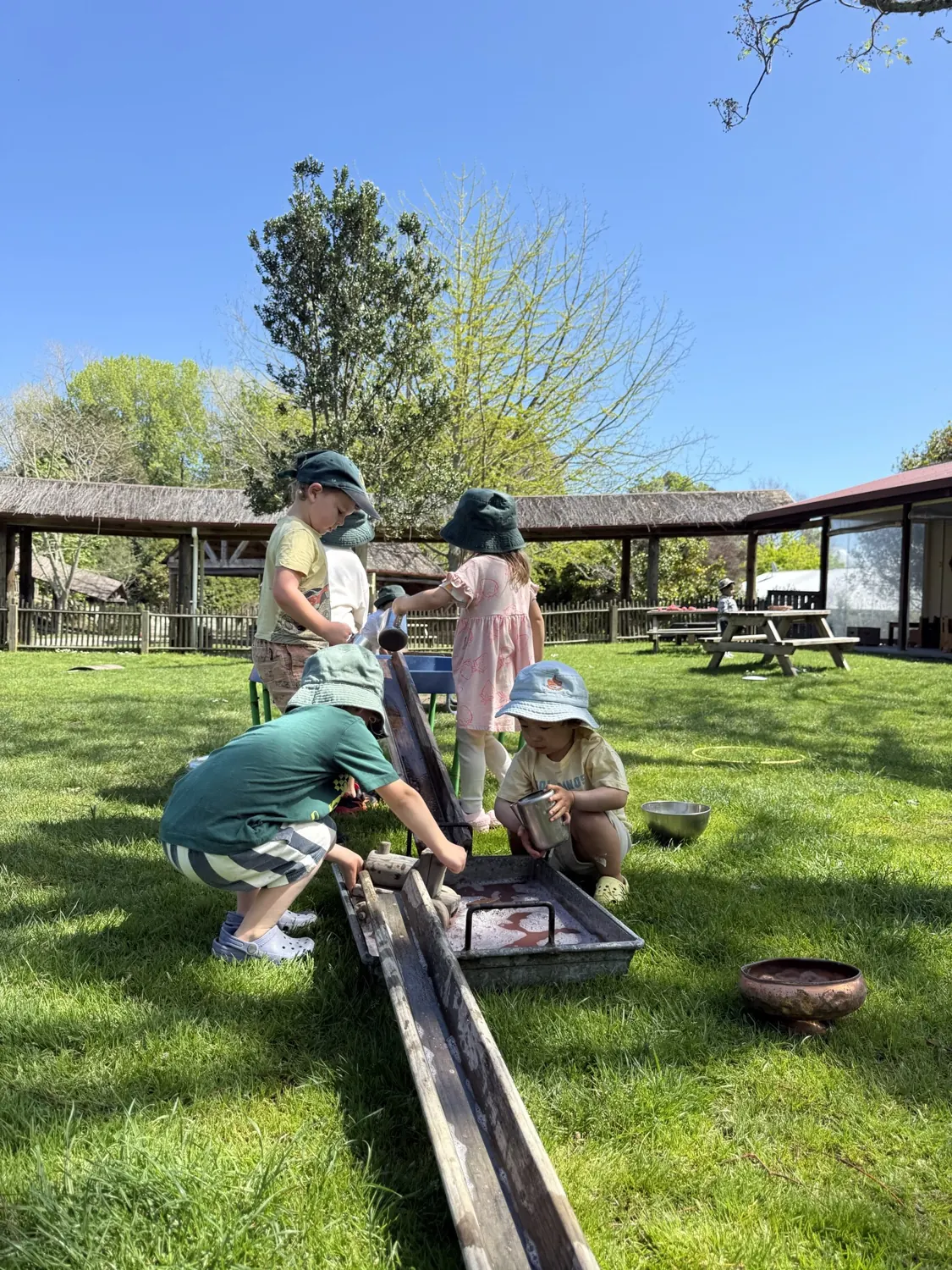 Children in sun hats playing at the water trough on the Tātua lawn