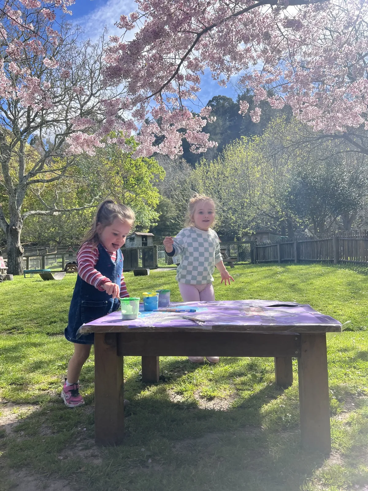 Two children painting at an outdoor table under the blossom tree, with The Farmyard lawns and trees behind them