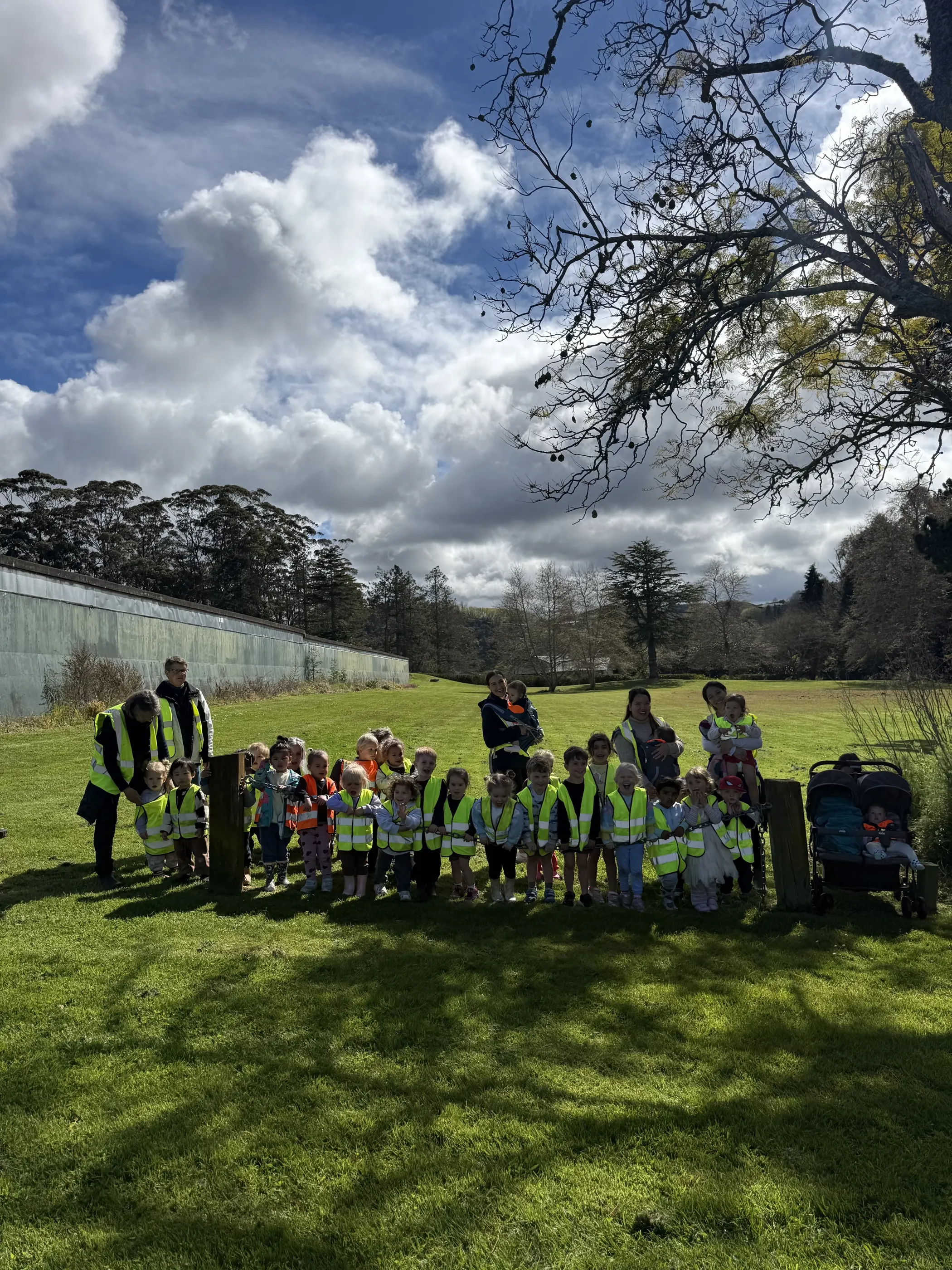Tamariki and kaiako in hi-vis vests gathered together on the big lawn under a wide sky